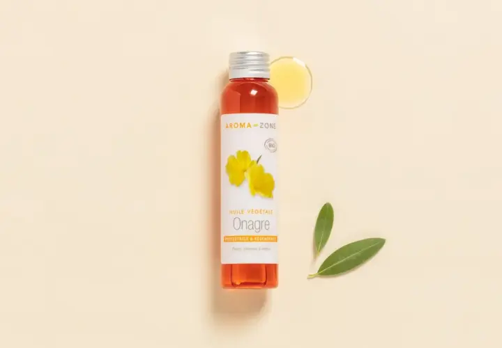 Flacon d'huile d'onagre Aroma-Zone, aux multiples bienfaits pour la peau et les cheveux. Une goutte d'huile dorée et deux feuilles vertes complètent la scène.