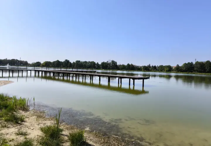 Drewniane molo w Krasnosielcu w letni, słoneczny dzień. Woda spokojna, odbija niebo i drzewa na horyzoncie.