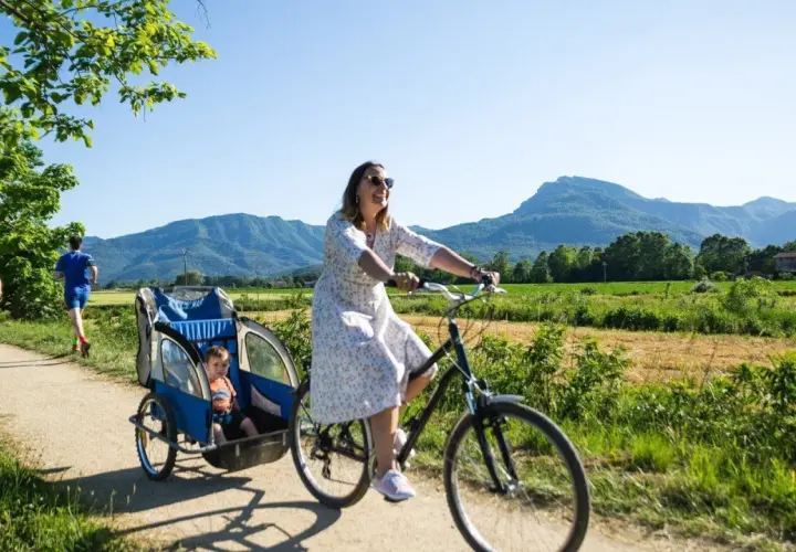 Disfrutando de un día soleado en la ruta el carrilet. Una mujer pedalea con un niño en un remolque, mientras otros ciclistas y un corredor disfrutan del paisaje montañoso.