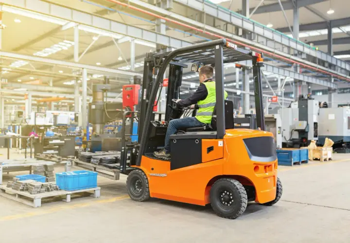 Ein Mann in Warnweste fährt einen orangefarbenen Gabelstapler in einer Fabrikhalle. Die Nutzungsdauer des Gabelstaplers ist entscheidend für die Effizienz.