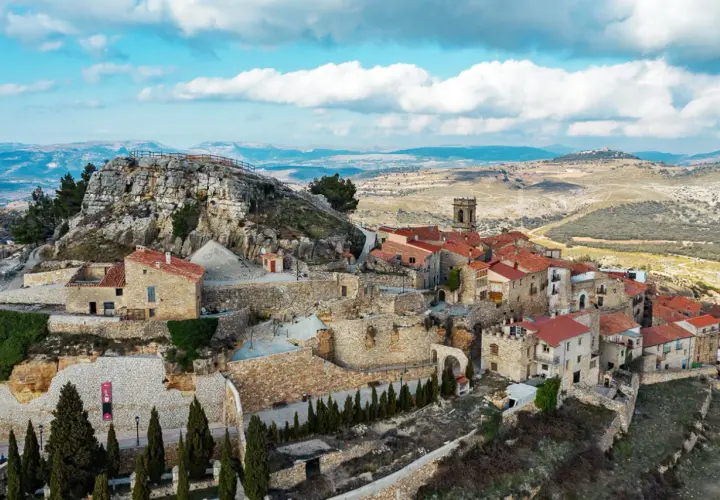 Vista aérea de un pueblo de casas de piedra con tejados rojos, anidado en la ladera de la roca del corb, bajo un cielo azul con nubes.