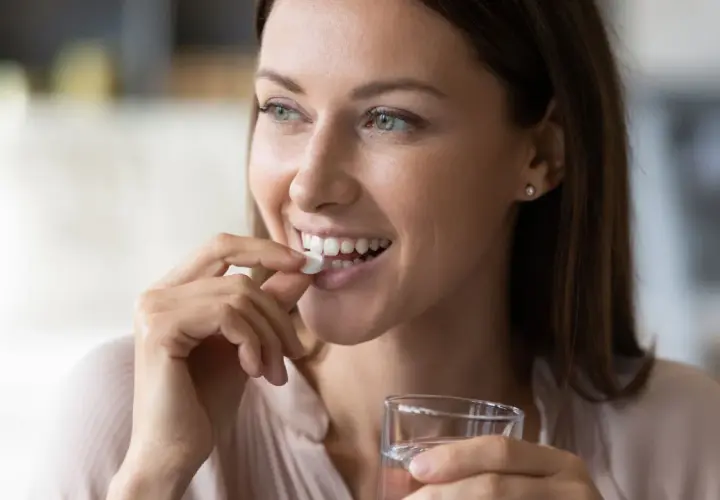 Femme souriante prenant un comprimé blanc, rappelant l'importance de ne pas associer le metronidazole alcool. Verre d'eau à la main.