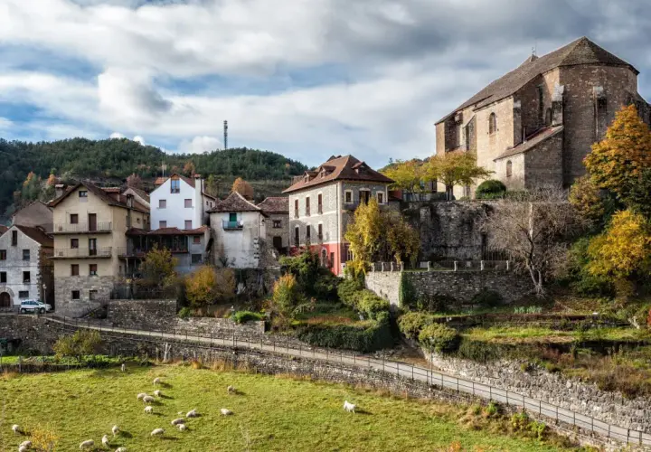 Casas de piedra y tejados rojos se asoman en el **valle de Ansó**, con ovejas pastando en un prado verde y un cielo nublado.