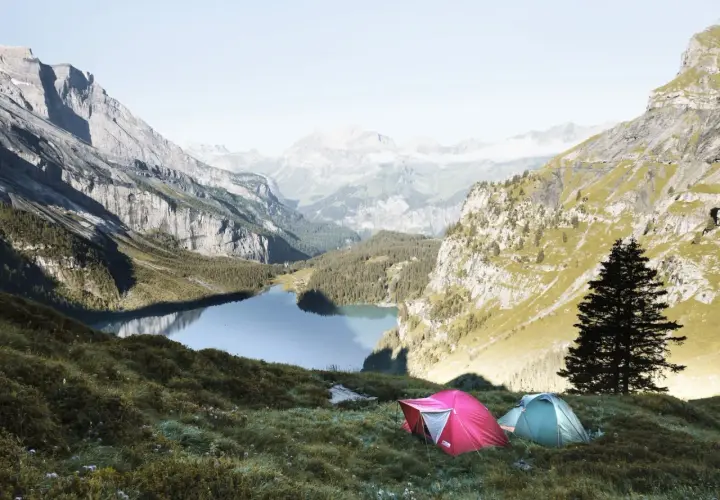 Two tents are set up on a grassy slope overlooking a serene lake in the Swiss Alps. This is a perfect spot for swiss alps hiking and camping.
