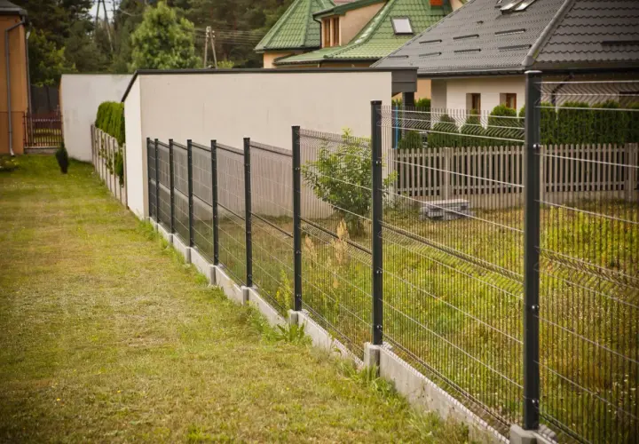 Nowoczesne ogrodzenie panelowe pod kątem wyznacza granicę posesji, za nim widać domy i zieleń.