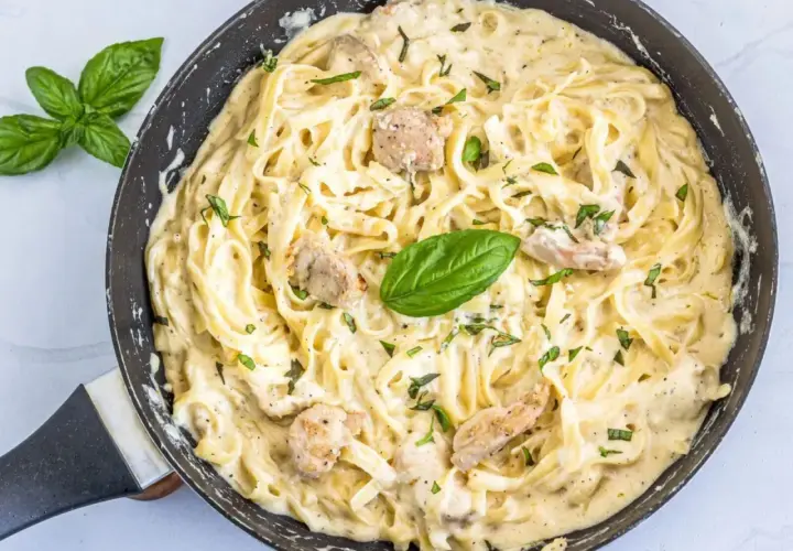 Un cremoso plato de pasta alfredo con trozos de pollo y hojas de albahaca fresca en una sartén.