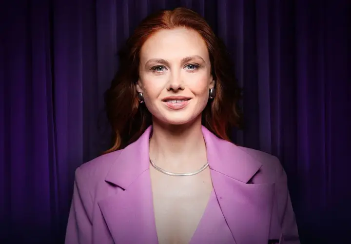 Anna Karczmarczyk w różowej marynarce, z rudymi włosami i niebieskimi oczami, uśmiecha się na tle fioletowej kurtyny. Jej filmy, seriale i programy cieszą się popularnością.