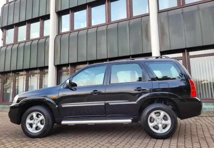 Czarny Mazda Tribute stoi na brukowanym podjeździe przed nowoczesnym budynkiem.