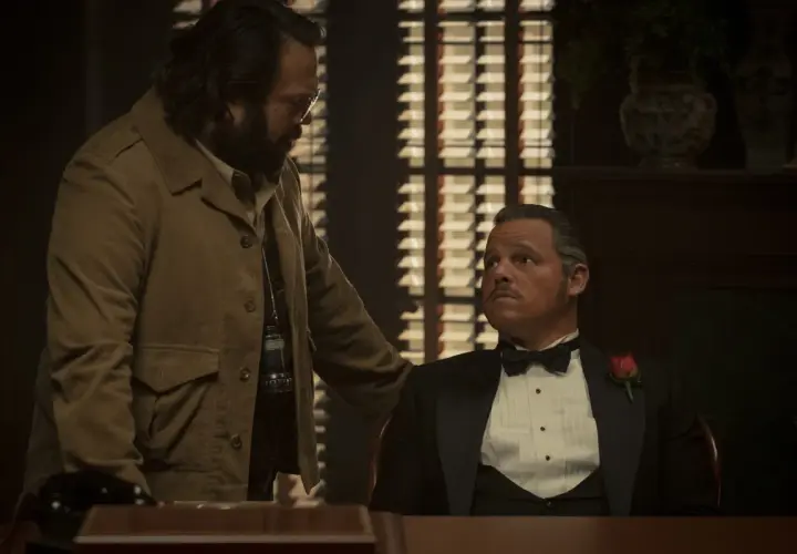 Un uomo con la barba e occhiali parla con un altro in smoking con una rosa. La scena suggerisce una recensione di un'offerta importante.