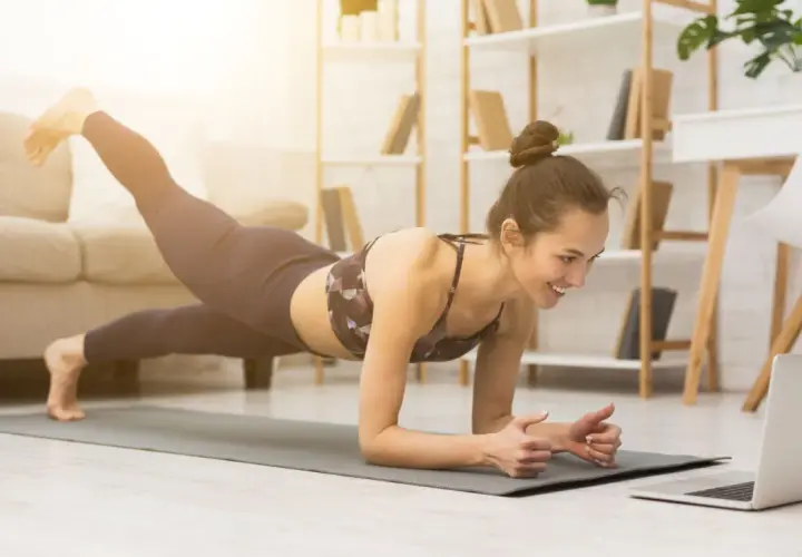 Uśmiechnięta kobieta w desce z nogą w górze, oglądająca trening na laptopie. To świetny sposób, jak zmotywować się do ćwiczeń w domu.