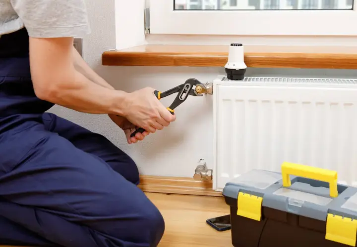 Instalator dokręca kluczem złączkę S do grzejnika, przygotowując go do sezonu grzewczego. Obok stoi skrzynka z narzędziami.