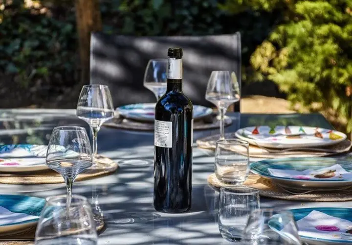 Une bouteille de vin rouge est prête pour un apéro dinatoire en extérieur, entourée de verres et d'assiettes sur une table de jardin.