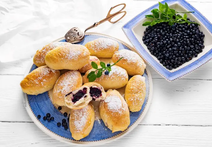 Pyszne jagodzianki posypane cukrem pudrem, obok miseczka świeżych jagód. Idealne na lato, zgodnie z najlepszym jagodzianki przepis.