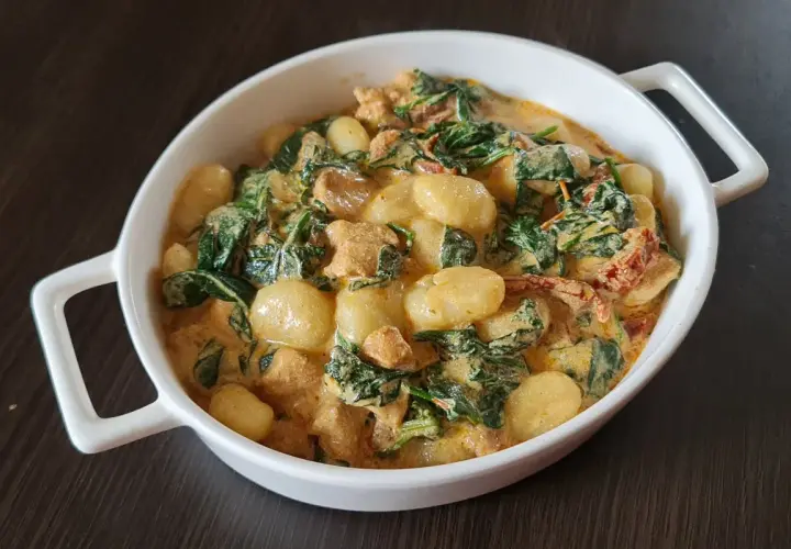Pyszne gnocchi z kurczakiem, szpinakiem i suszonymi pomidorami w kremowym sosie. Danie podane w białej owalnej misce.
