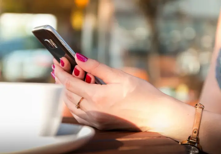 Dłoń z pomalowanymi paznokciami trzyma smartfon, obok biała filiżanka. Długą żywotność baterii w telefonie doceni każda użytkowniczka.