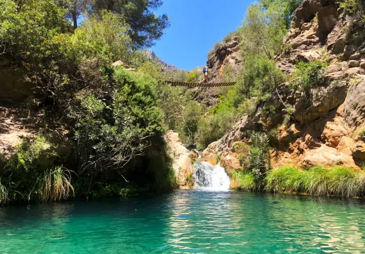 Un puente colgante cruza un **río verde** en Granada, con una cascada y vegetación exuberante.