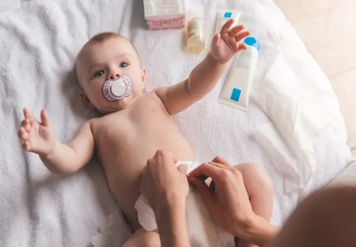 Chłopiec z smoczkiem leży na ręczniku, a ręce mamy dokonują **przewijania chłopca**. Obok kosmetyki i chusteczki.