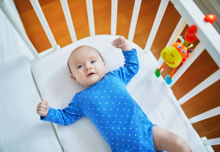 Urocze, 10 tygodniowe dziecko w niebieskim pajacyku leży w łóżeczku obok kolorowej zabawki.