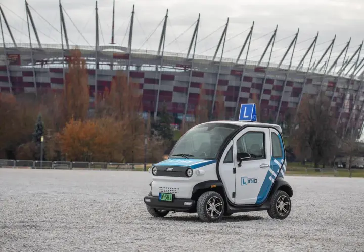 Mały biały samochód z niebieskimi pasami i literą "L" na dachu, na tle stadionu.