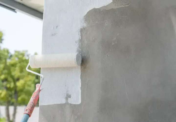 Malowanie betonu wałkiem. Biała farba pokrywa szarą ścianę, odświeżając jej wygląd.