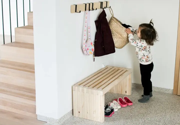 Dziecko wiesza torbę na haczyku nad drewnianą ławką. Prosty projekt, jak zrobic ławkę, idealny do przedpokoju.