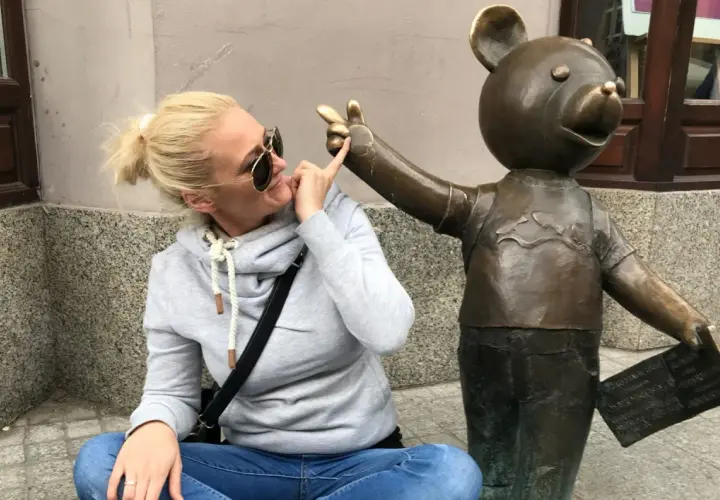 Kobieta w okularach przeciwsłonecznych i szarej bluzie bawi się z pomnikiem misia, który trzyma tabliczkę. To część szlaku łodzi bajkowej.
