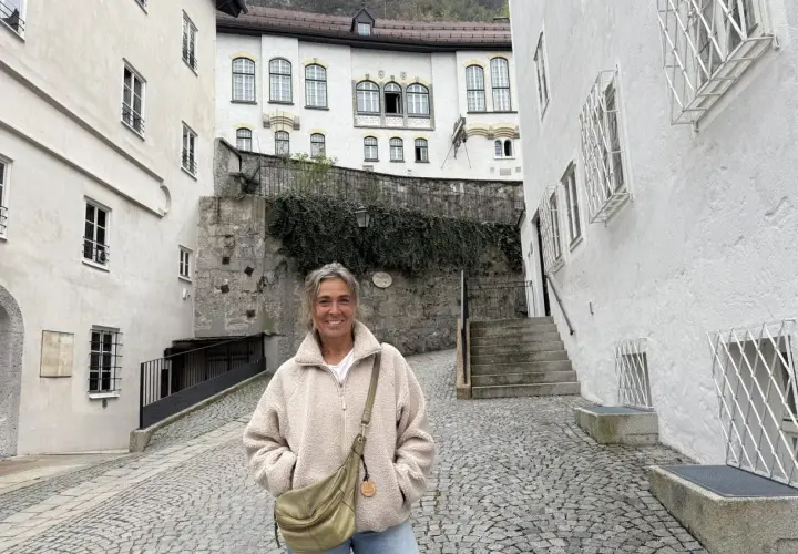 Eine Frau steht lächelnd auf einem Kopfsteinpflasterweg zwischen alten weißen Häusern. Sie könnte eine Pilgerin sein, die auf ihrer Reise neue Orte entdeckt.