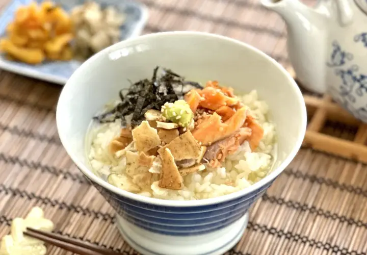 A delicious ochazuke recipe featuring rice, flaked salmon, nori, and wasabi in a blue-striped bowl, with a teapot in the background.