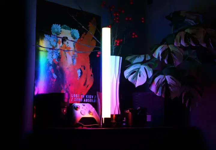 Na półce stoją kontroler do gier, aparat i lampa, której światło RGB tworzy nastrojową atmosferę.