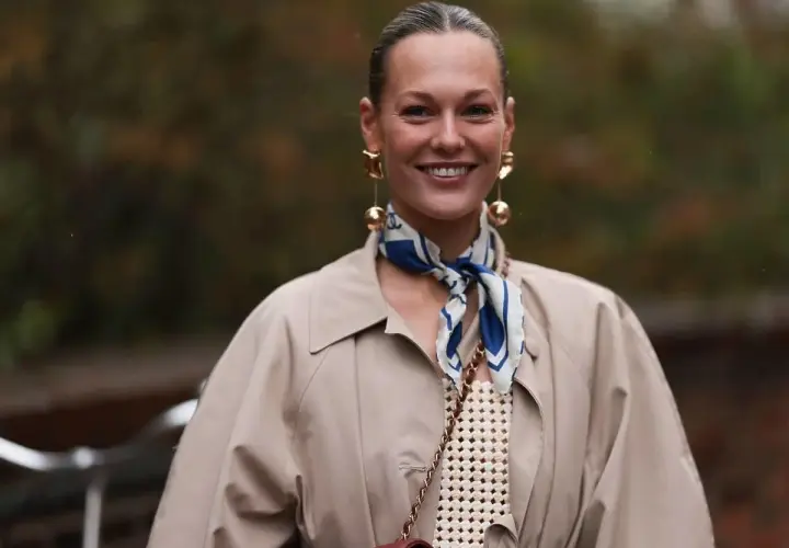Uśmiechnięta kobieta w beżowym płaszczu i stylowej apaszce z niebieskimi wzorami. Inspiracja, jak nosić apaszkę, by dodać elegancji.