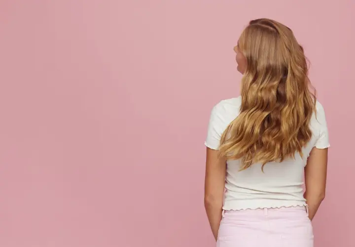 Blond włosy z efektem butterfly cut, falujące i lśniące, na tle pudrowego różu.