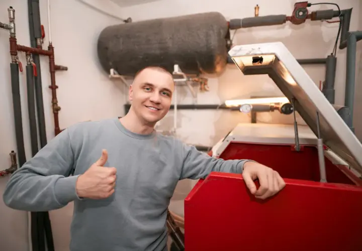 Uśmiechnięty mężczyzna pokazuje kciuk w górę obok czerwonego kotła i dużego zbiornika wyrównawczego CO.