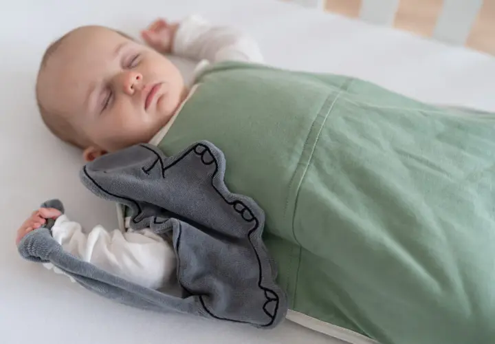 A baby sleeps soundly in a green sleep sack, a grey elephant toy peeking out. This image inspires thoughts on how to make baby clothes.