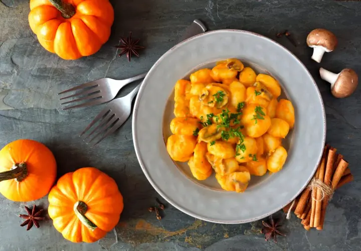 Pyszne gnocchi z kremowym sosem dyniowym, ozdobione natką pietruszki. Obok talerza małe dynie i przyprawy.