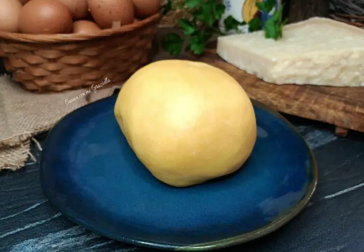 Impasto giallo per frolla salata su piatto blu, con uova e parmigiano sullo sfondo.