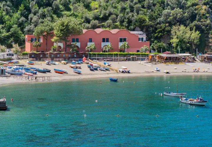 Plage de Vico Equense avec des bateaux colorés sur le sable et dans l'eau turquoise. Bâtiment rose en arrière-plan.