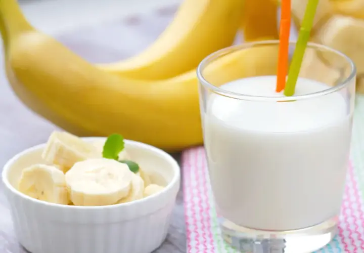 Szklanka pysznego mleka bananowego z dwoma słomkami, obok miseczka z pokrojonymi bananami i całe banany w tle.