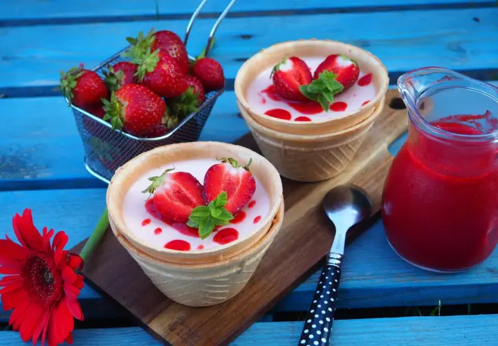 Panna cotta w wafelkach z truskawkami i miętą, obok dzbanuszek z musem truskawkowym.