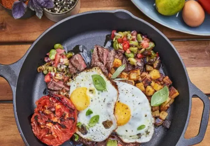 Un plat de petit-déjeuner copieux dans une poêle en fonte : steak grillé, deux œufs au plat, pommes de terre rôties, tomate grillée et une salade de légumes. Un vrai oeuf barbecue !