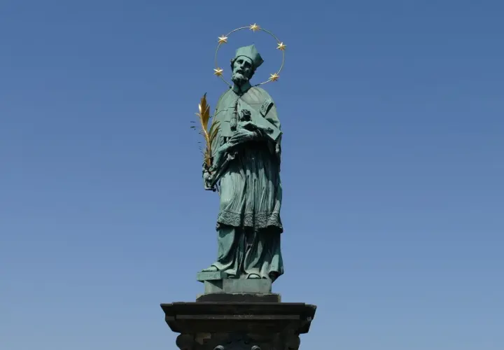 Pomnik św. Jana Nepomucena z gwiazdami nad głową i palmą męczeństwa w dłoni, na tle błękitnego nieba.