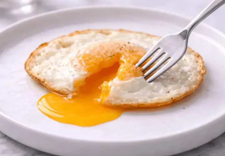 Un œuf miroir, le jaune coulant comme un soleil liquide, est percé par une fourchette sur une assiette blanche.