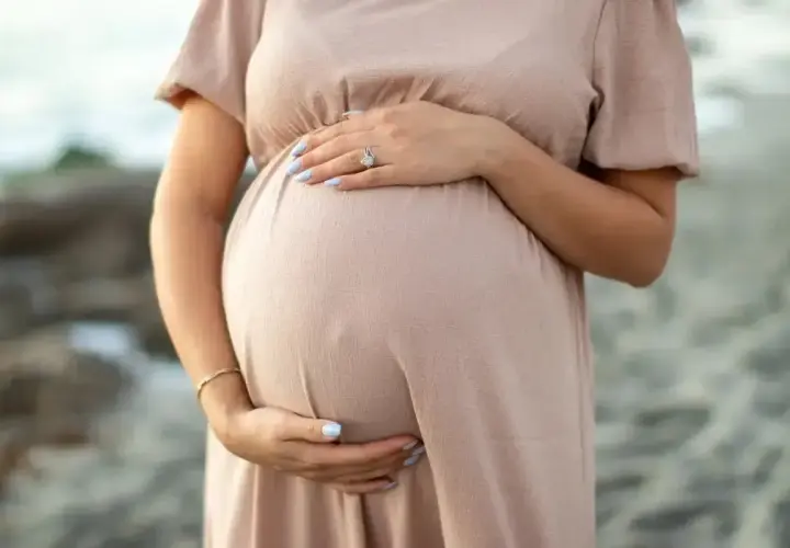 Brzuch kobiety w ciąży, delikatnie otulony beżową sukienką. Dłonie z jasnoniebieskim lakierem na paznokciach spoczywają na brzuchu. Warto pamiętać o bezpieczeństwie, np. unikając retinolu w ciąży.