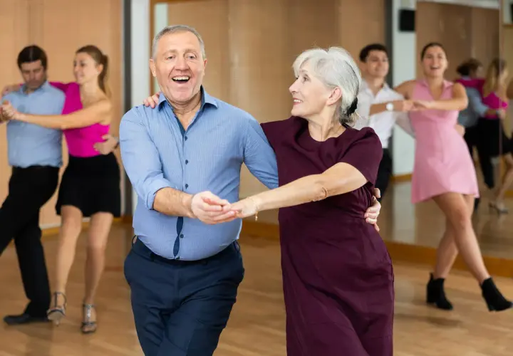 Uśmiechnięta para seniorów w rytmie tańca na 3. W tle inne pary ćwiczą kroki.