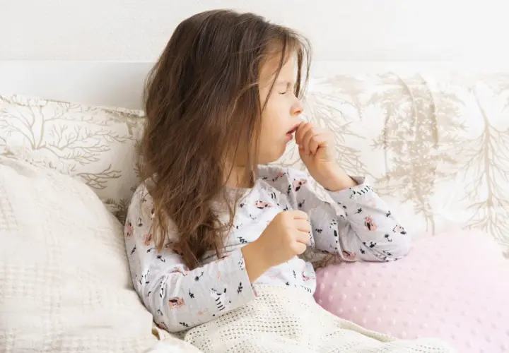 Bambina con tosse stizzosa, coperta da una coperta, seduta a letto con cuscini decorati.