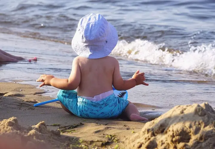 Niemowlę w kapeluszu i kąpielówkach z rekinami siedzi na plaży, ciesząc się wakacjami z niemowlakiem nad morzem.