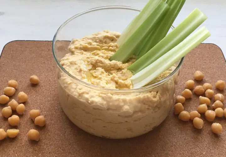 Domowy hummus przepis: kremowa pasta z ciecierzycy w szklanej misce, podana z łodygami selera i ziarnami ciecierzycy.