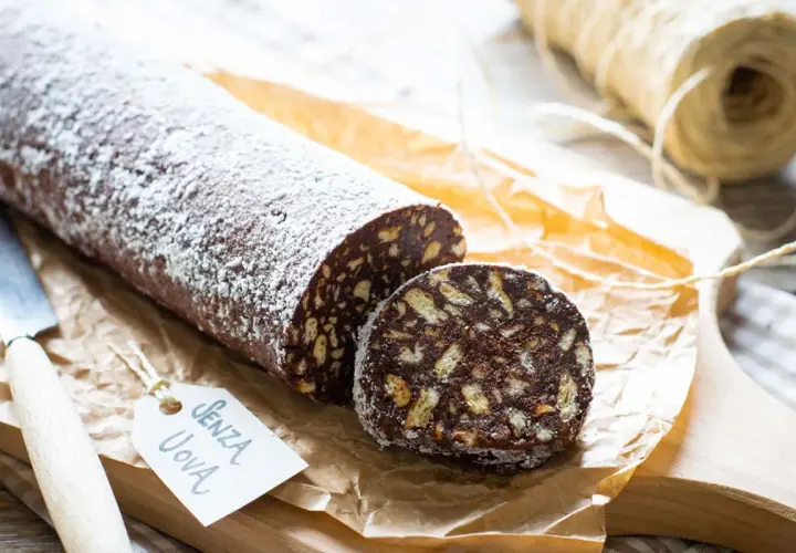 Salame al cioccolato senza uova, cosparso di zucchero a velo, con una fetta tagliata che mostra i biscotti sbriciolati.