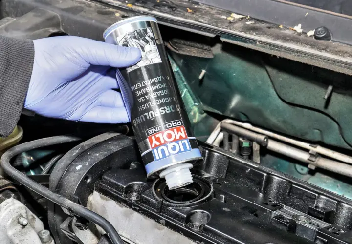 Ręka w niebieskiej rękawiczce wlewa Liqui Moly płukanie silnika do otworu w pokrywie zaworów.