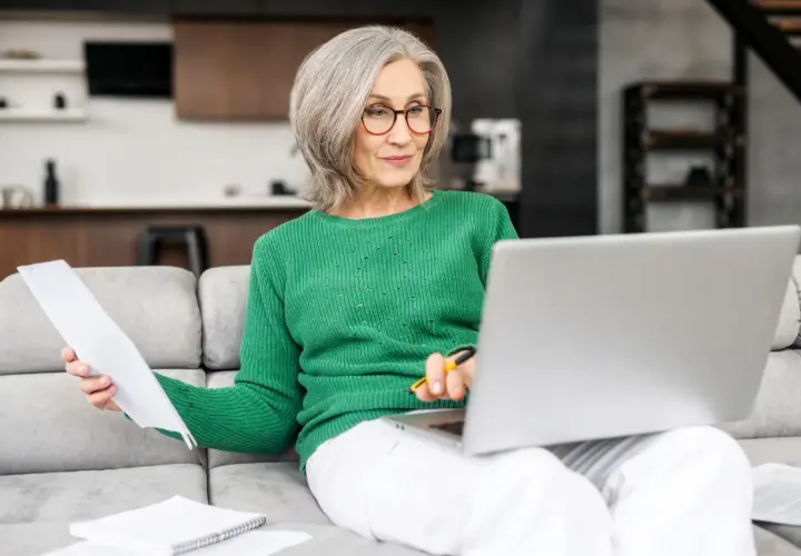 Femme âgée souriante, assise sur un canapé, travaillant sur son ordinateur portable et consultant des documents. Elle semble gérer son **apa à domicile montant**.
