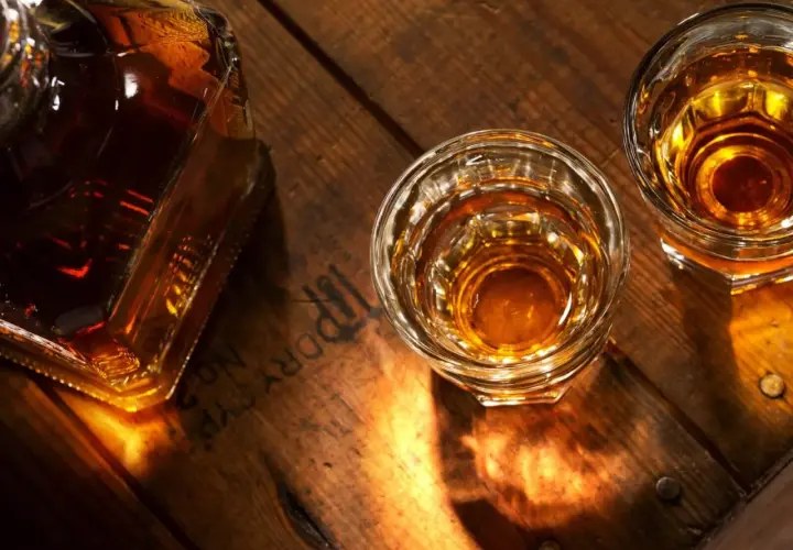 Butelka i dwa kieliszki z bursztynowym płynem, przypominającym brandy lub whisky, stoją na drewnianym blacie.